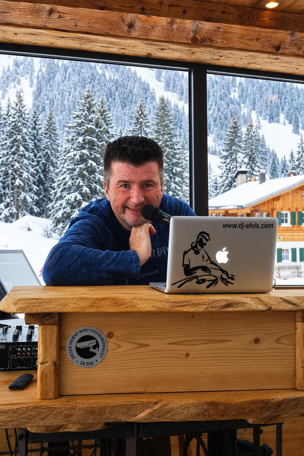 DJ Elvis mit Mikrofon und Laptop am Holzpult vor verschneiter Alpenlandschaft