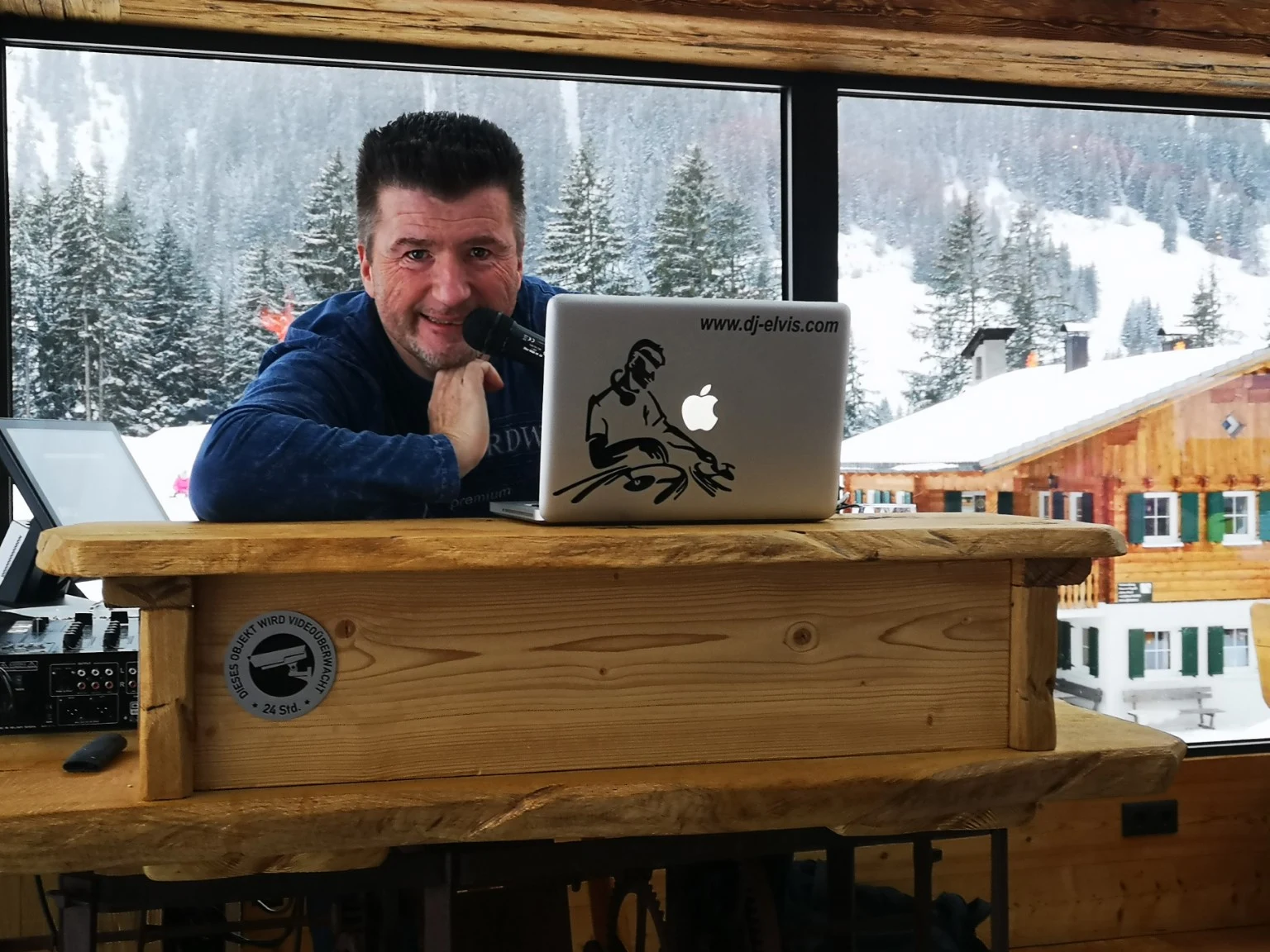 Originalfoto von DJ Elvis mit Mikrofon am Holzpult vor verschneiten Tannen